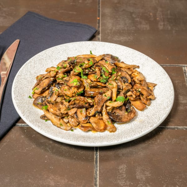 A plate of sautéed mushrooms with fresh parsley, a knife, and a dark napkin on a tiled surface.