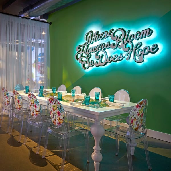 A long dining table with clear chairs, teal glasses, and a neon sign on a green wall.