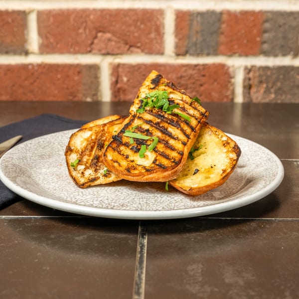 Grilled bread slices with char marks and fresh parsley on a white plate, against a brick wall.