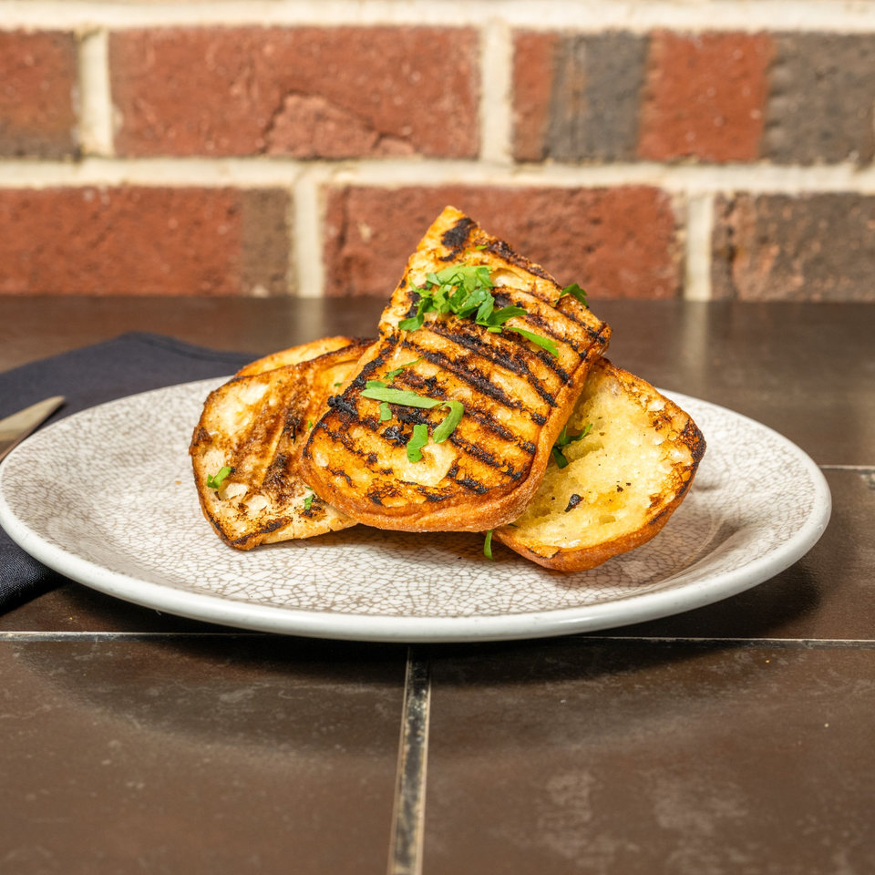 Grilled bread slices with char marks and fresh parsley on a white plate, against a brick wall.