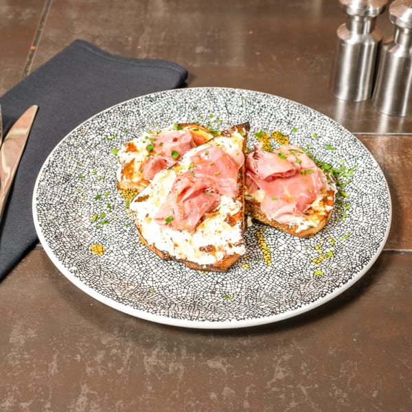 Three pieces of Lena's bruschetta topped with prosciutto, creamy cheese, and fresh herbs on a patterned plate.
