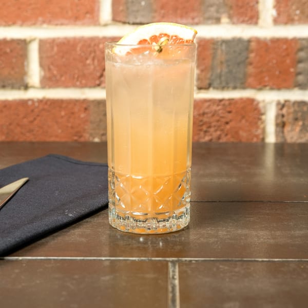A tall, clear glass of a light orange cocktail with a grapefruit slice, on a dark table.