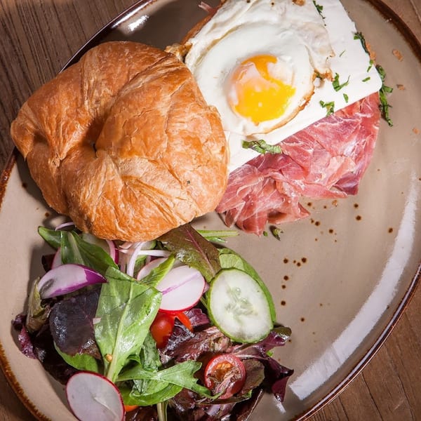 Croissant with prosciutto, fried egg, and a side salad with radishes and cucumbers on a plate.