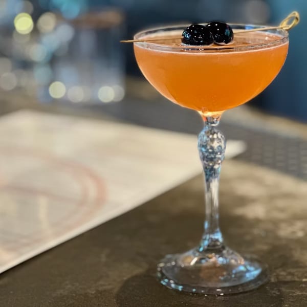 A vibrant orange cocktail in a coupe glass, garnished with two dark cherries on a toothpick.