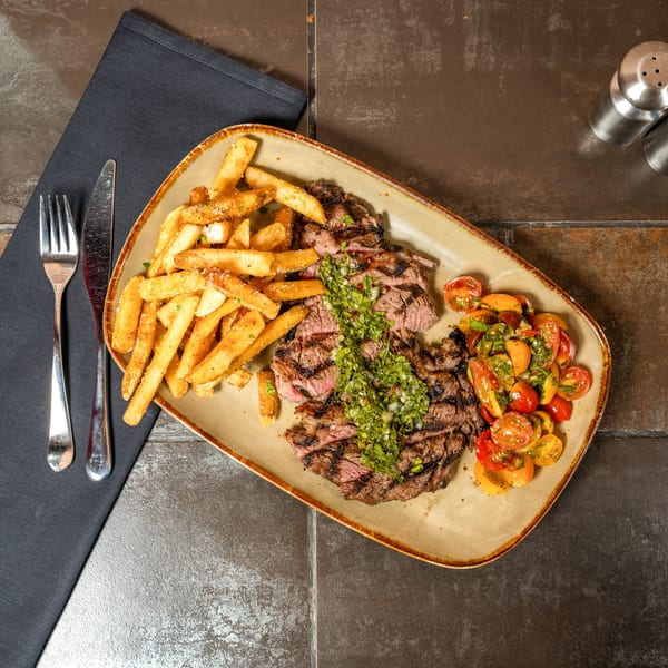 Grilled steak with chimichurri, a side of fries, and cherry tomato salad on a plate.