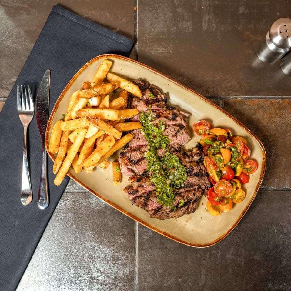Grilled steak with chimichurri, a side of fries, and cherry tomato salad on a plate.