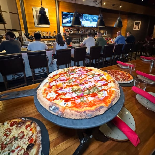 Three pizzas on a wooden table with a bar and patrons in the background at Lena's.