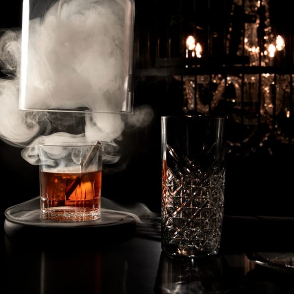 Smoked cocktail with a cinnamon stick, next to an empty patterned glass, with dim lighting.