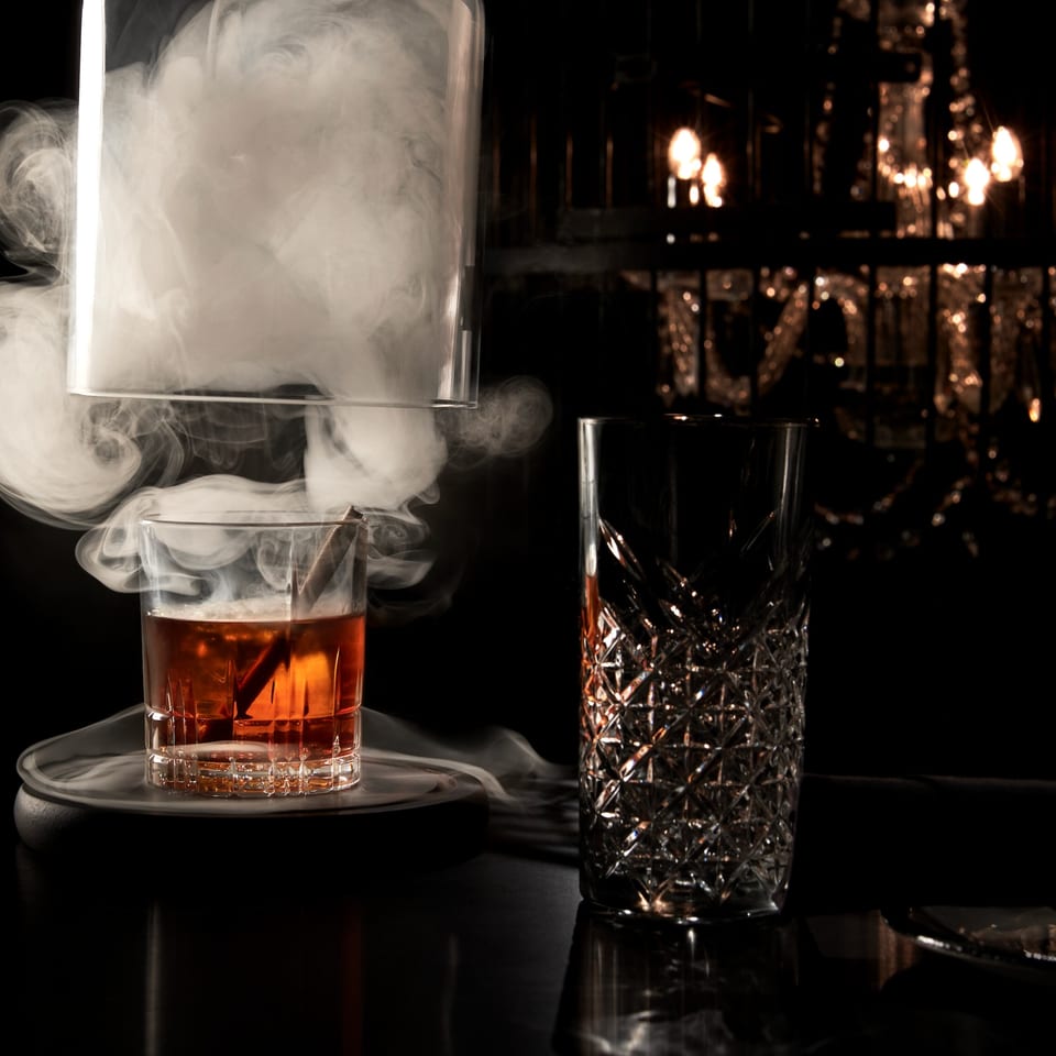 Smoked cocktail with a cinnamon stick, next to an empty patterned glass, with dim lighting.