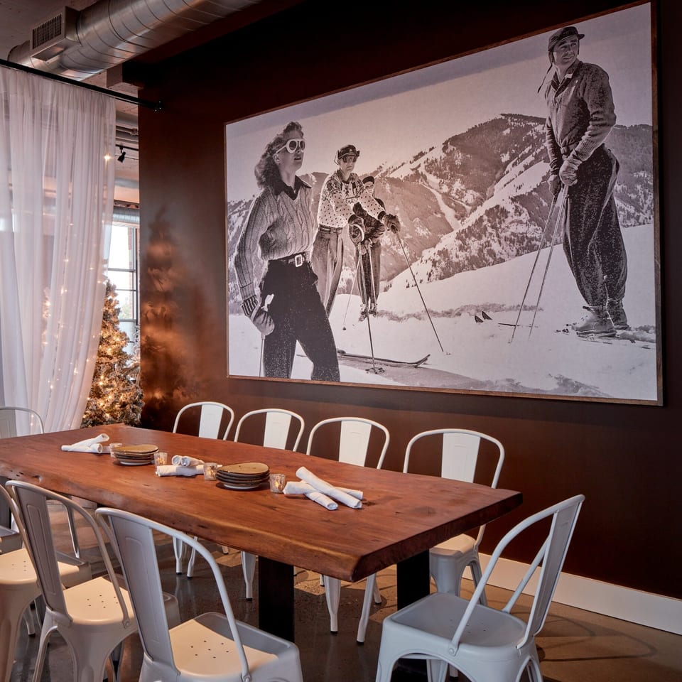 Long wooden table with white chairs, set with plates and napkins, next to a large ski-themed mural.