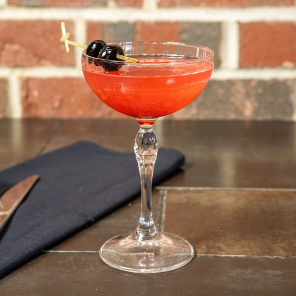 Red cocktail in a coupe glass, garnished with two dark cherries on a skewer, against a brick wall.