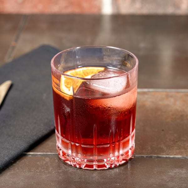 A vibrant red Negroni cocktail with ice and an orange slice, on a dark table.