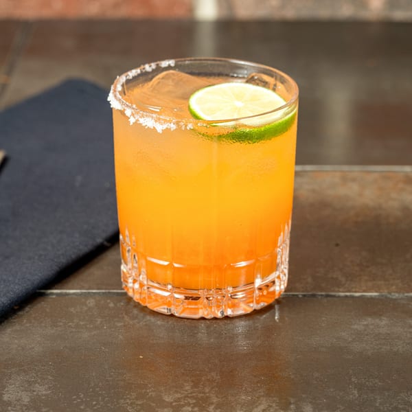 A vibrant orange cocktail with a salted rim and lime slice sits on a dark table.