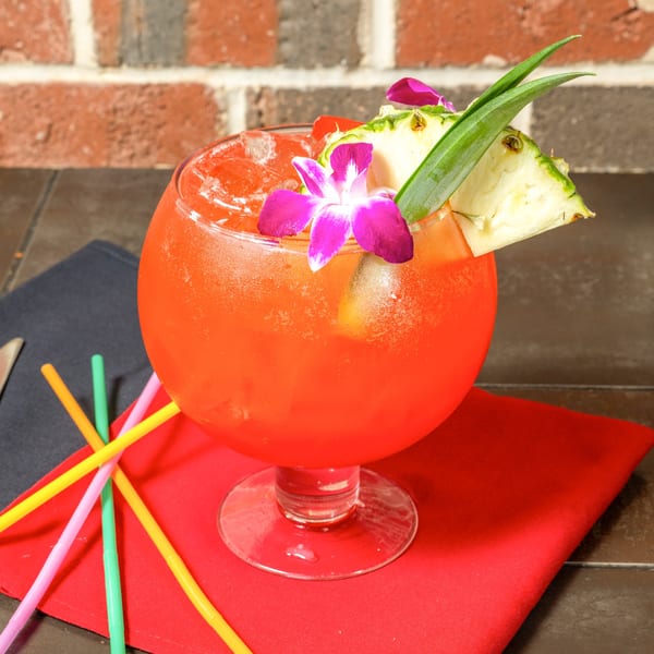 A vibrant red cocktail in a large glass, garnished with pineapple, a cherry, and an orchid.