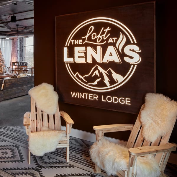 A glowing "The Loft at Lena's Winter Lodge" sign above two wooden chairs with white fur throws.