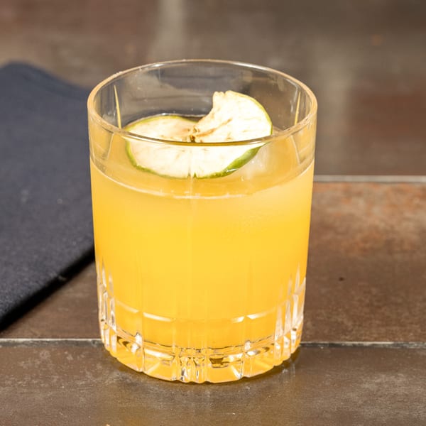 A vibrant orange cocktail in a cut glass, garnished with a dried apple slice, on a dark table.