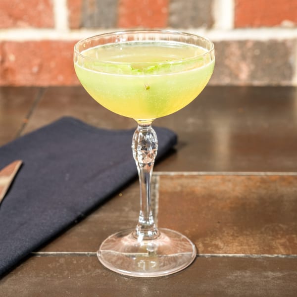 A vibrant green cocktail in a coupe glass with a decorative stem, on a dark table with a brick wall.