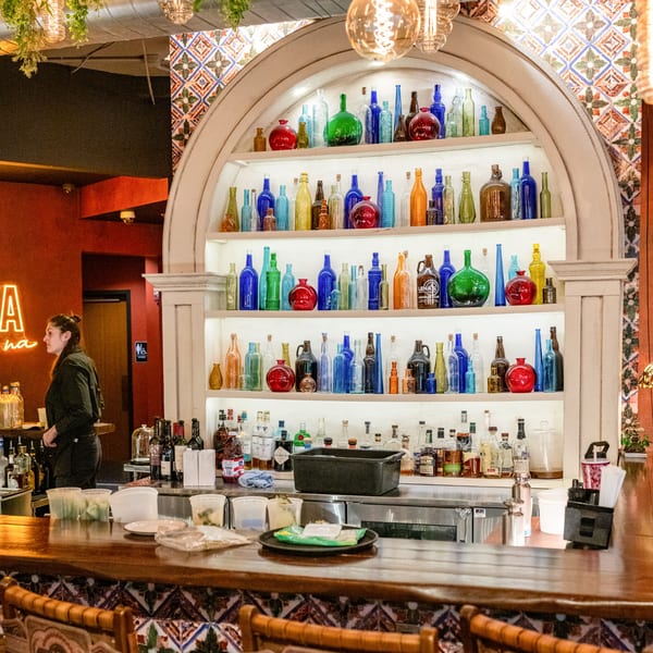 A bartender stands behind a wooden bar with colorful bottles displayed on shelves at Lena's Wood-Fired Pizza.