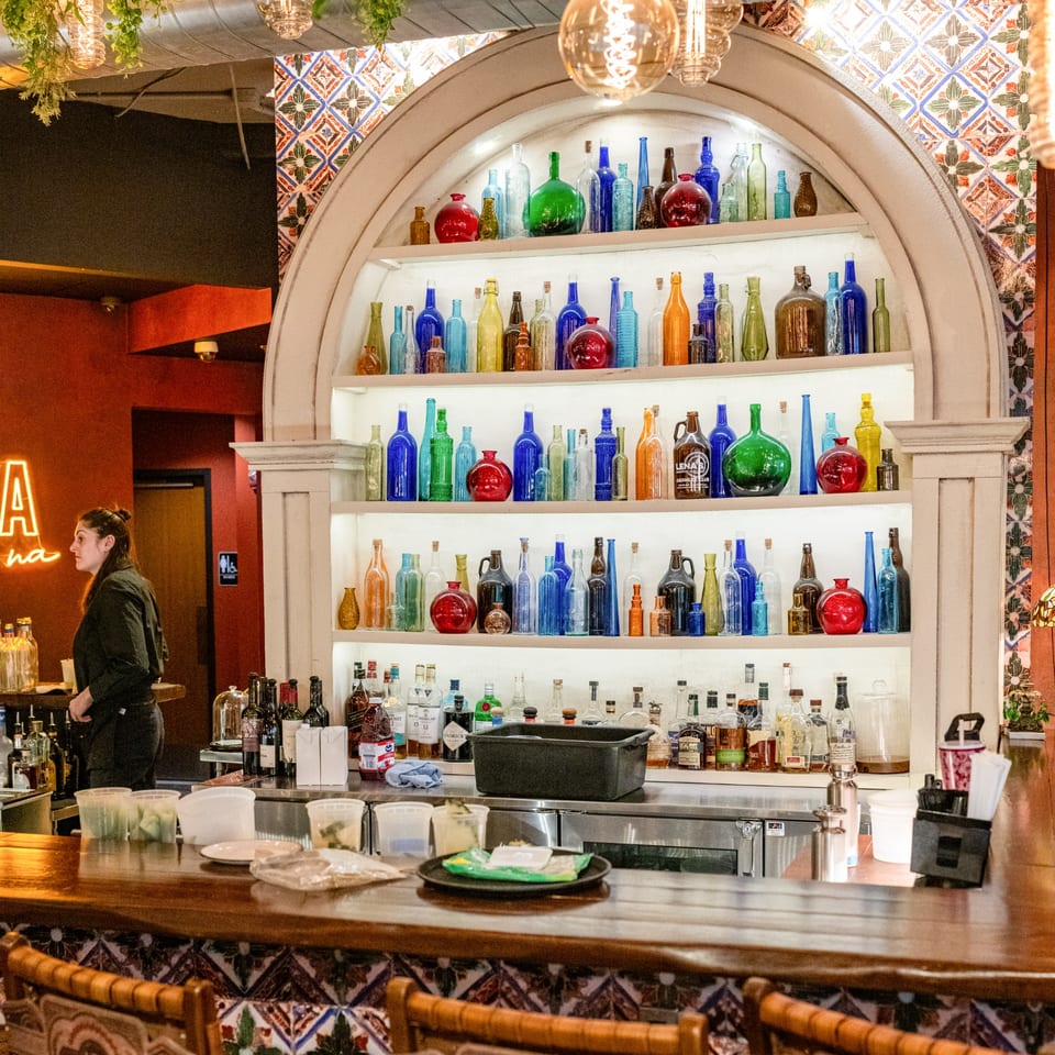 A bartender stands behind a wooden bar with colorful bottles displayed on shelves at Lena's Wood-Fired Pizza.