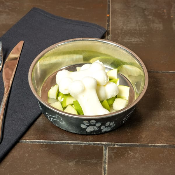 Dog bowl with bone-shaped treats and chopped green apples on a dark tiled surface.