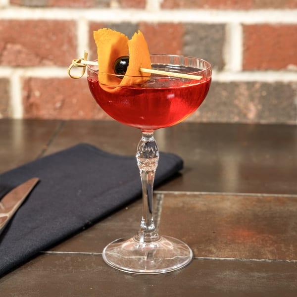 A red cocktail in a coupe glass, garnished with an orange peel and cherry, on a dark table.