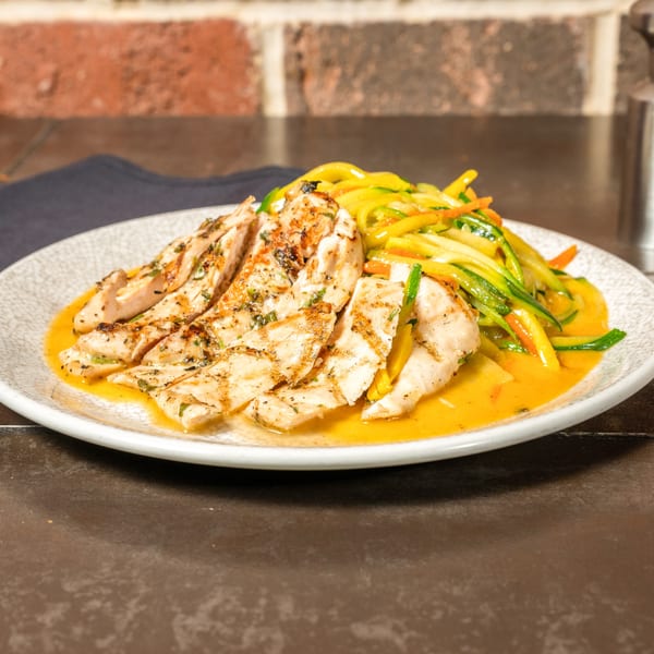 Grilled chicken slices with zucchini noodles and carrots in a yellow sauce on a white plate.