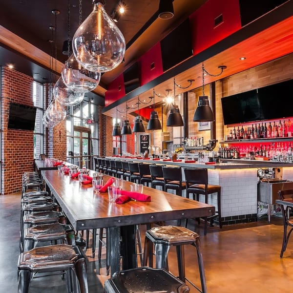 Long communal table with stools and a full bar with red lighting at Lena's Wood-Fired Pizza & Tap.