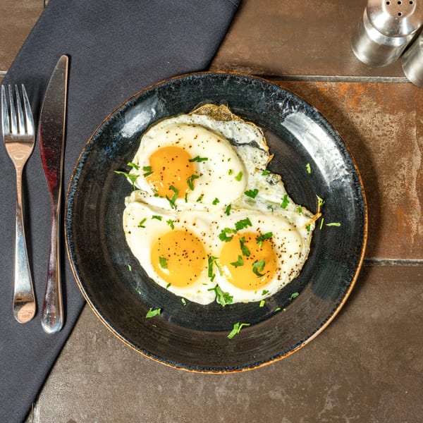 Three sunny-side-up eggs with parsley and pepper on a dark plate, with cutlery and napkin.
