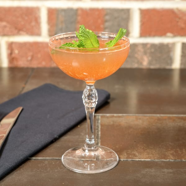 A peach-colored cocktail with mint garnish in a coupe glass on a dark table with a brick wall.