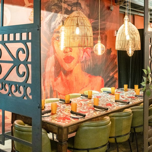 Long dining table with green chairs, orange glasses, and a colorful patterned top, under wicker pendant lights.