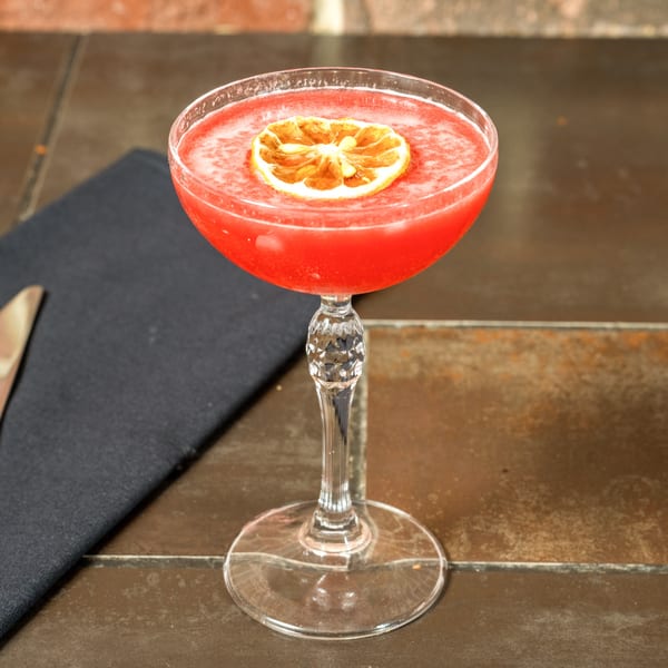 A vibrant red cocktail in a coupe glass, garnished with a dried orange slice, on a dark table.