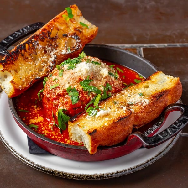 Lena's Wood-Fired Pizza & Tap's large meatball in red sauce, topped with cheese and parsley, served with toasted bread.