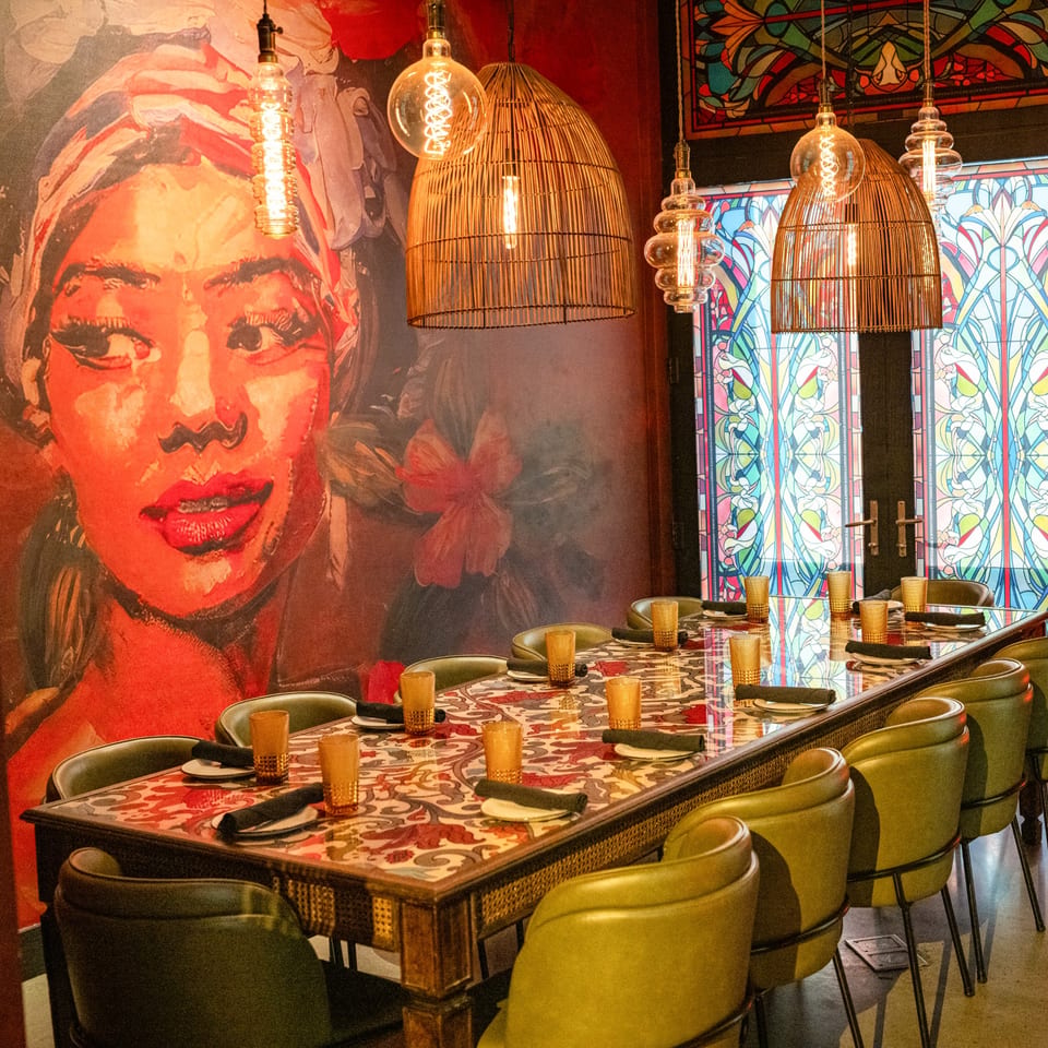 Long dining table with green chairs, patterned top, and a vibrant mural of a woman on the wall.