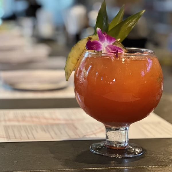 A vibrant red cocktail in a goblet, garnished with pineapple, green leaves, and a purple orchid.