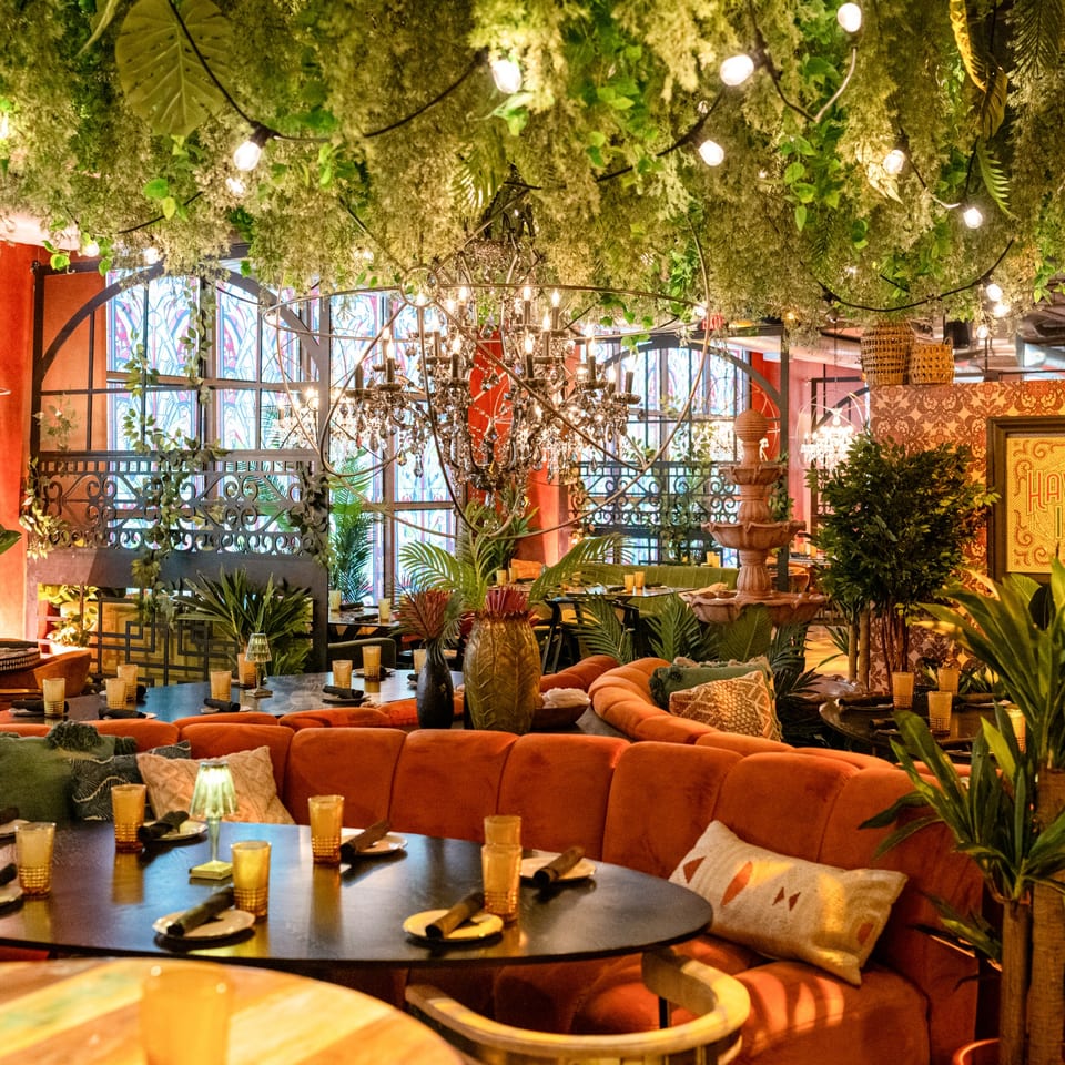 Lena's Wood-Fired Pizza & Tap dining room with orange banquettes, lush greenery, and chandeliers.