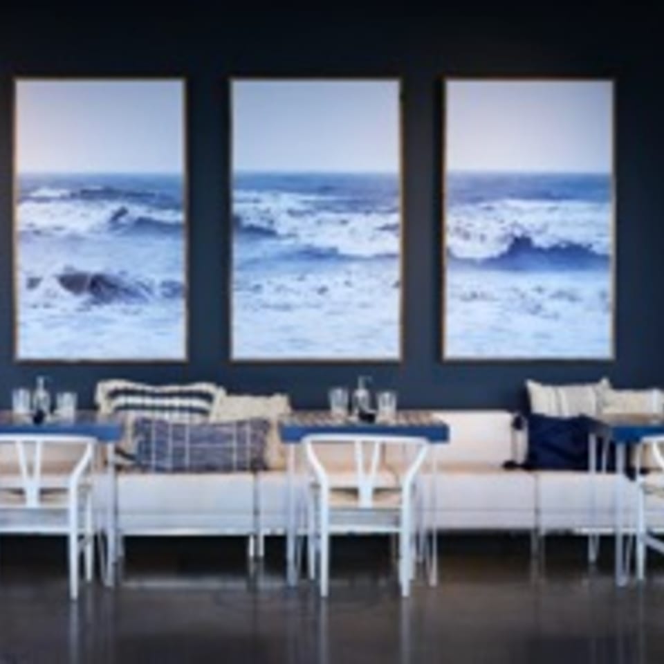 Restaurant interior with blue walls, three ocean wave paintings, white benches, and blue tables.